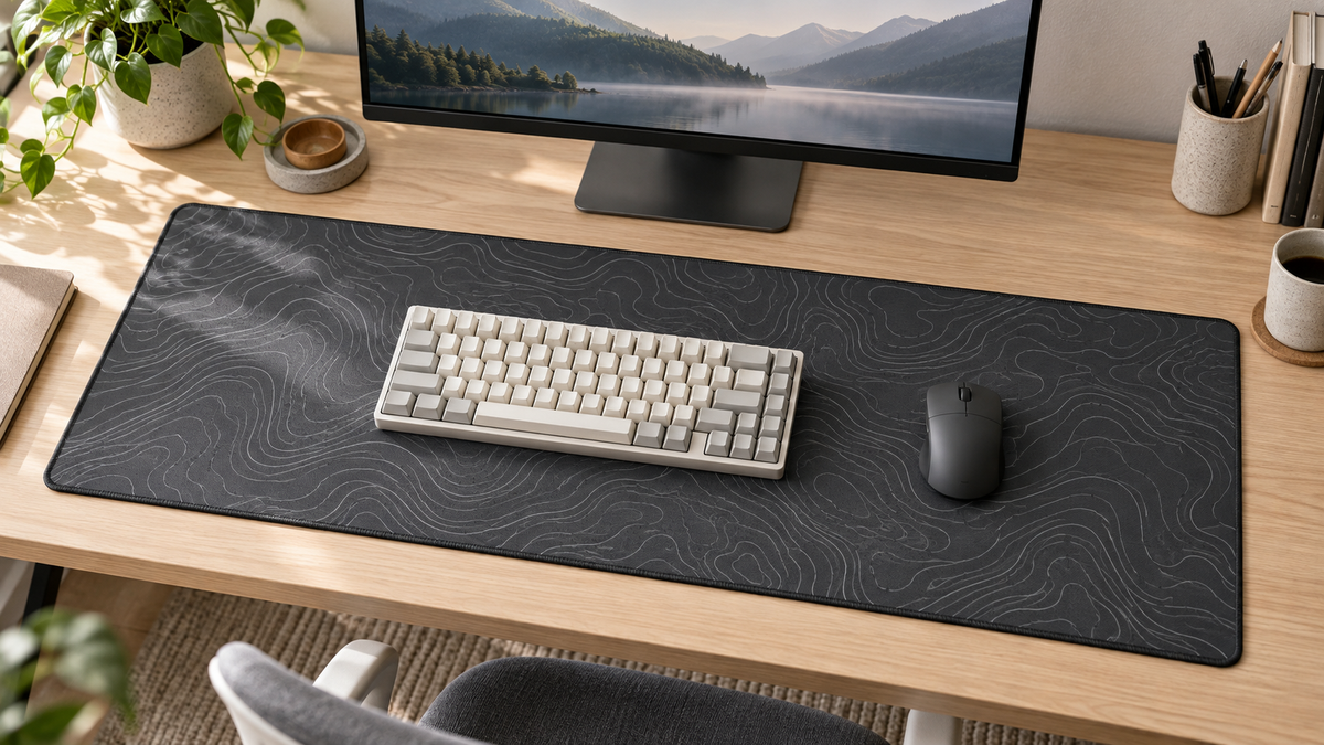 Illustrative minimalist desk with a charcoal contour-print fabric mat, compact cream keyboard and simple planter.
