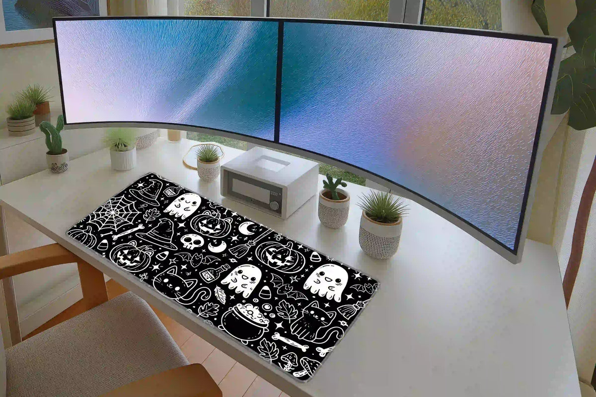 Black and white Halloween doodle mat beneath two monitors, with cheerful ghosts, pumpkins and cats across the desk