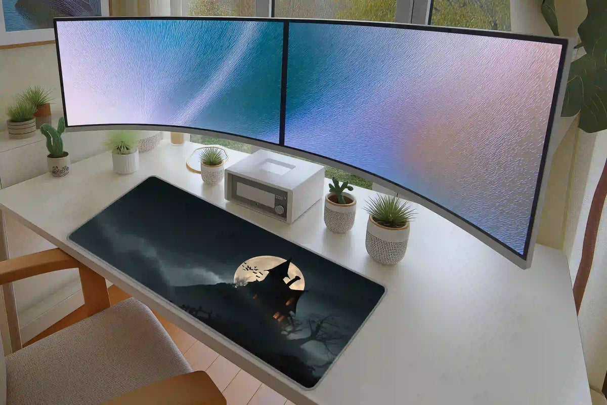 Haunted house desk mat on a white desk below curved monitors, showing the moonlit mansion and dark misty sky