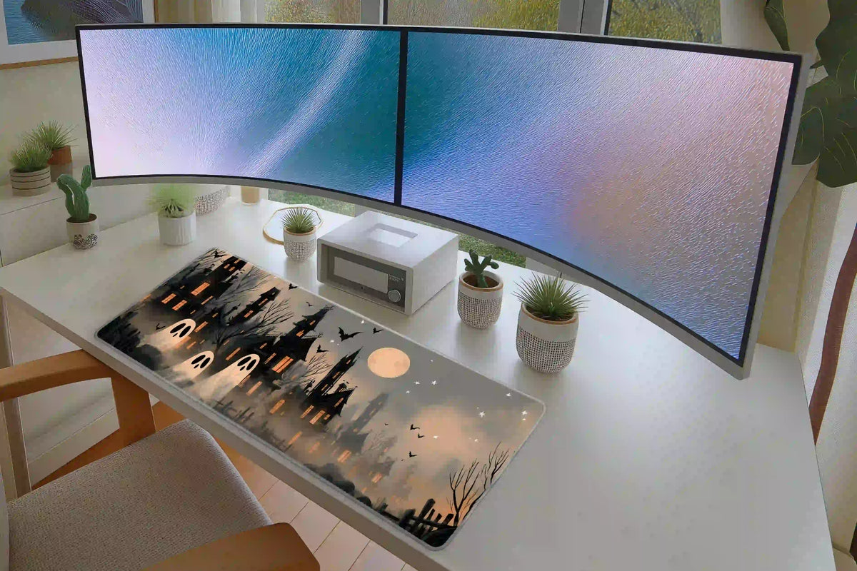Haunted village desk mat with cream ghosts and black houses on a white desk beneath two curved monitors